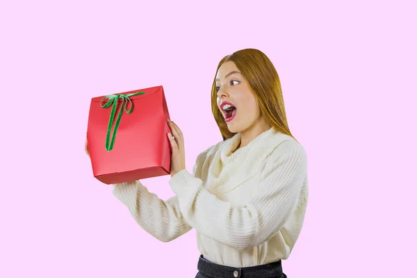Piękna śliczna ruda dziewczyna, która trzyma worek na prezent pudełko czerwone z zieloną aksamitną wstążką podnosząc prezent pokazuje jego uśmiechnięty entuzjastycznie patrząc na bok przed izolowanym — Zdjęcie stockowe