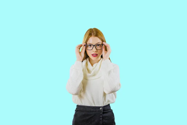 Ένα χαριτωμένο νεαρό κοκκινομάλλα κορίτσι με τα γυαλιά κρατά ένα χείλος των γυαλιών με τα δύο χέρια και με μια αυστηρή ματιά με το κεφάλι της κάτω κοιτάζοντας κάτω στην κάμερα σε ένα απομονωμένο φόντο — Φωτογραφία Αρχείου