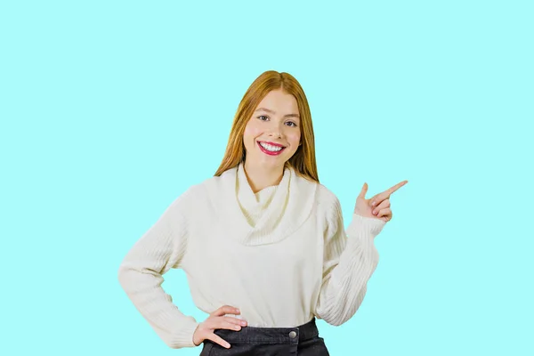 Ritratto di una giovane ragazza dai capelli rossi che mostra il dito indice di lato e sorride guardando la fotocamera su uno sfondo isolato — Foto Stock