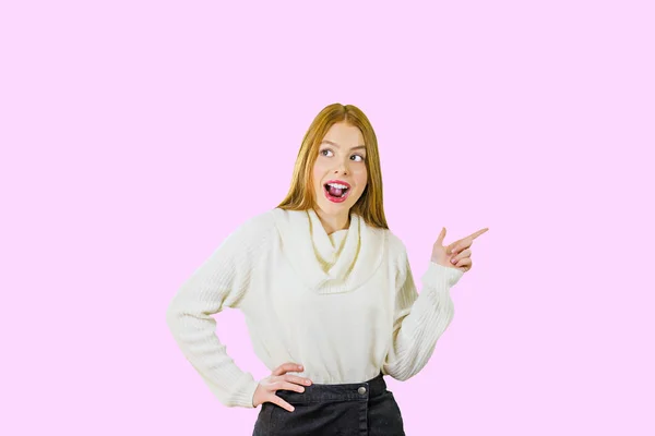 Πορτρέτο ενός νεαρού κοκκινομάλλη κορίτσι που δείχνει το δείκτη της στο πλάι και χαμογελά ενώ κοιτάζει στο αντίθετο του δείκτη του χεριού σε ένα απομονωμένο φόντο — Φωτογραφία Αρχείου