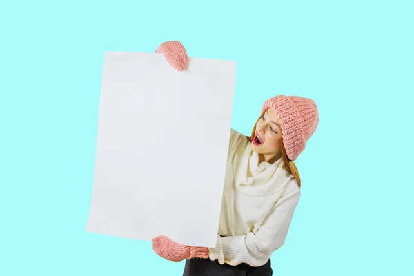 A young red-haired girl in a knitted pink hat and mittens holds a large white poster with both hands and looks at the poster with her mouth wide open on an isolated background — Stock Photo, Image