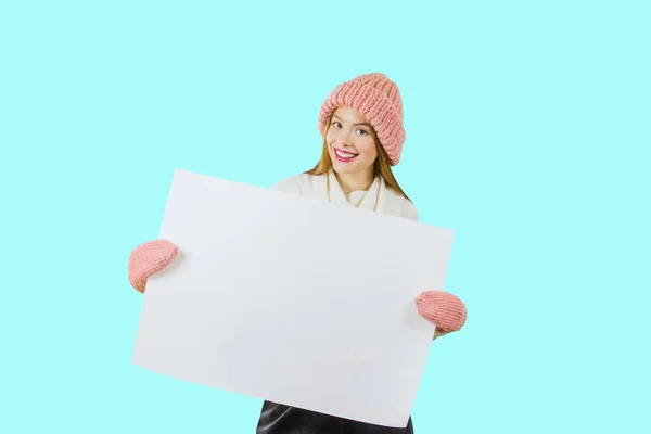 Portrait of a young red-haired girl in a knitted pink hat holding a poster with both hands closing herself with a poster and looking at the camera against an isolated background — Stock Photo, Image