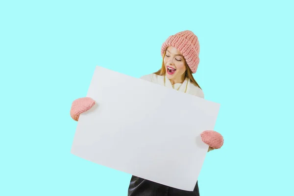 Seorang gadis berambut merah muda dengan topi rajutan merah muda memegang poster dengan kedua tangan menutupi dirinya dengan poster dan melihat poster dengan mulut terbuka di latar belakang yang terisolasi — Stok Foto