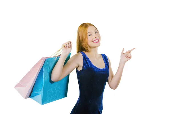 Een schattig roodharige meisje in een blauw fluwelen jurk met zakken over haar schouder shows met haar wijsvinger aan de zijkant en kijkt in de camera terwijl glimlachend — Stockfoto