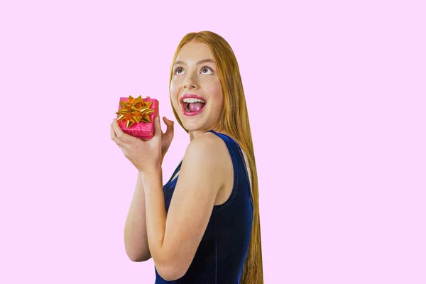 Attraente giovane ragazza dai capelli rossi con un regalo in mano vicino al viso sorride su uno sfondo rosa isolato — Foto Stock