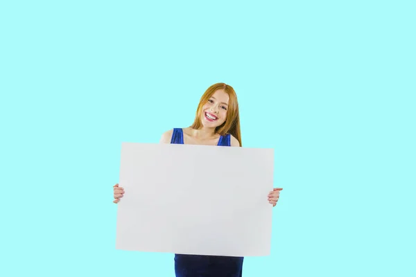 Portrait of a young red-haired girl who is holding a large white poster or piece of paper posing at the camera and smiling, showing white teeth on an isolated background — Stock Photo, Image