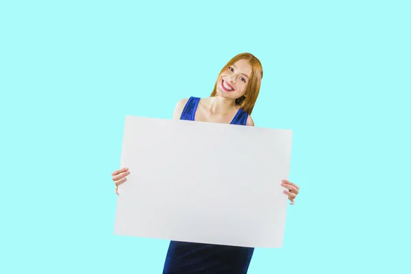 Una giovane ragazza dai capelli rossi sta tenendo un grande poster bianco o un pezzo di carta in posa alla fotocamera con un sorriso su uno sfondo isolato — Foto Stock