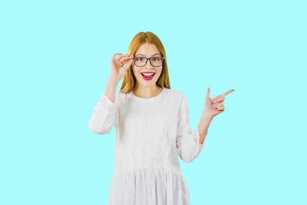 Portrait of a young beautiful red-haired girl in glasses, which shows the emotions of enthusiastic joy, looking into the camera, showing the index finger to the side, photo on an isolated background — Stock Photo, Image