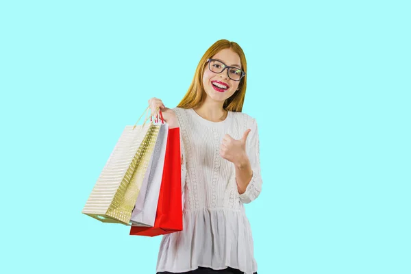 Een portret van een jong roodharige meisje in een witte blouse met gekleurde vakantie tassen die haar handen vasthouden met een hand, terwijl de andere hand toont de klas en glimlacht naar de camera — Stockfoto