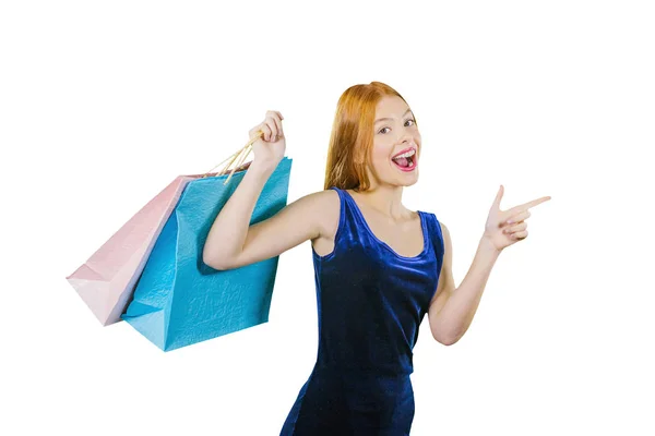 A beautiful young red-haired girl in a dress holds three packages, raising her hand with the packages, and with her other hand shows her index finger to the side and looks into the camera while Royalty Free Stock Photos