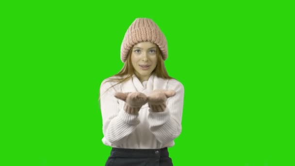 Buon Natale e Felice Anno Nuovo vacanze piene di divertimento. La ragazza in un cappello a maglia rosa e guanti imita la neve nelle sue mani, soffiandola dalle sue mani. Sorridente ragazza felice su sfondo verde — Video Stock