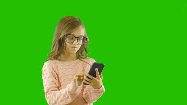Seorang gadis kecil yang cantik dari usia sekolah membaca dan mencari sesuatu di ponselnya, menemukan dan dengan tulus bersukacita dengan menggenggam tangannya, menembaki latar belakang yang terisolasi — Stok Video