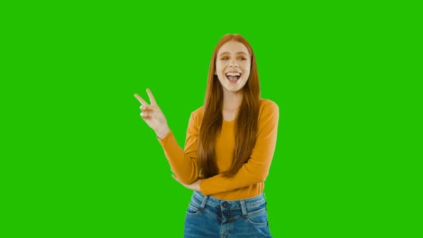 明るいメイクアップの感情的な笑顔を持つ魅力的な赤毛の女の子は、感情的に笑顔を示し、緑の背景に2本の指を上げて片手で示しています — ストック動画
