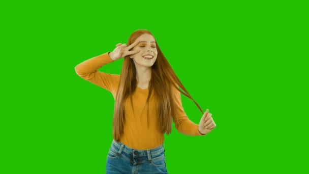 Cheerful attractive young red-haired girl in a playful mood, holding two fingers of one hand near her eye, and the second takes her long hair, green background — Stock Video