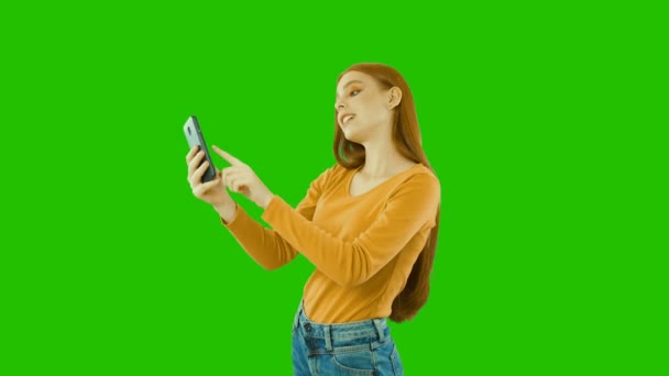 Attractive pretty red-haired girl looking at her smartphone, typing sms, watching the news and showing her positive emotions, green background — Stock Video