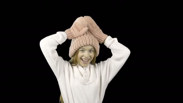 Une jeune adolescente en bonnet rose tricoté et mitaines roses se livre à montrer ses cornes avec ses mains et souriant, vacances d'hiver, tournage sur un fond isolé — Video