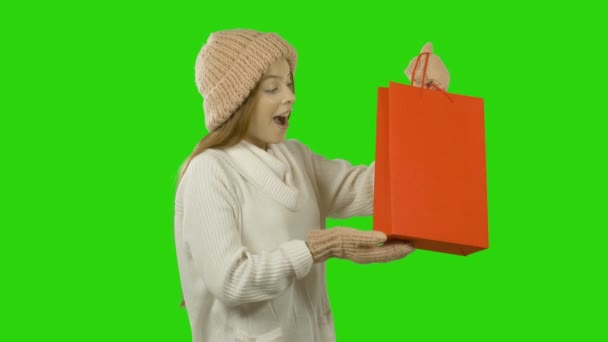 Una giovane ragazza con cappello e bambole tiene un pacchetto con un regalo rosso, lo guarda e sorride, vacanze invernali, umore natalizio, sfondo verde — Video Stock