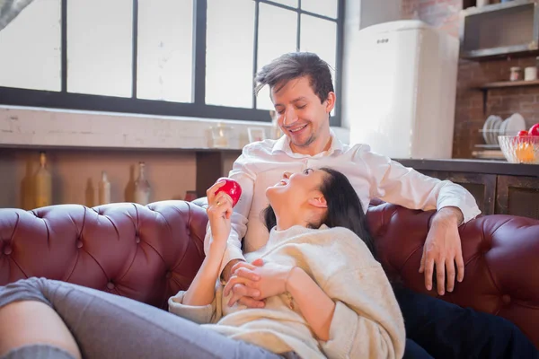 A Beautiful Joyful Girl in an embrace looks at a Guy, looks at him with an apple in his hands on a sofa in the kitchen — Stock Photo, Image