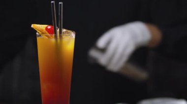 Tekila gündoğumu kokteylini yavaşça masaya yatırın. Barmen elleri arka planda çalkalayıcı çalıyor. Güzel bir kadeh uzun içki. Bar içkileri serisi.