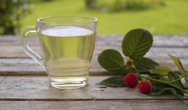 Dışarıdaki eski ahşap masada şeffaf bir fincan ahududu yaprağı gelgiti yakın çekim. Masada taze çiğ ahududu var. Bitkisel tıp konsepti