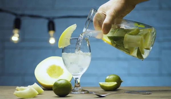 Kavun, kireç, nane yaprağı ve buzla dolu bir testiden bir bardağa karpuz limonatası dolduran erkek elini kapat. Mavi arkaplanda bulanık lambalar