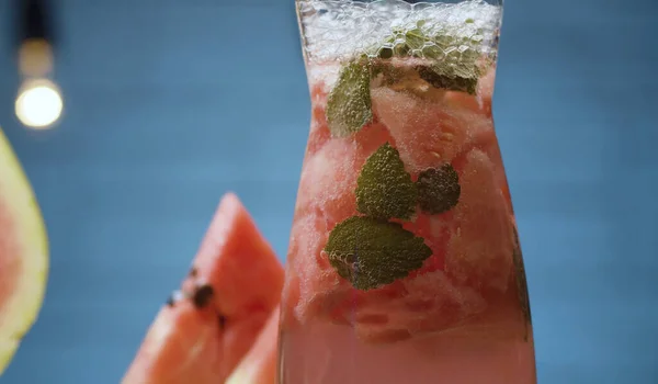 Karpuz limonatasını cam sürahide kapat. Soda, karpuz parçaları, nane yaprakları ve cam sürahide buz. Mavi arkaplanda bulanık lambalar