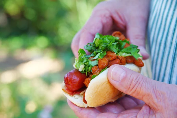 Chili dog with fresh chopped lettuce held by older man in apron in a backyard