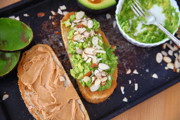 Overhead of sandwich preparation with rye bread, peanut butter, mashed avocado and sliced almonds