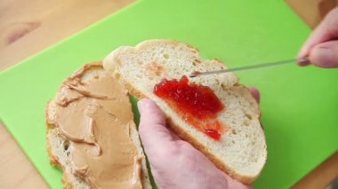 Bir adam yeşil kesme tahtası nın üzerine fıstık ezmeli ve reçelli sandviç yapar.