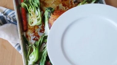 Maşa kullanarak, bir kadın beyaz bir tabak üzerine bir kızarmış tavuk çeyrek, bebek bok choy ve havuç koyar