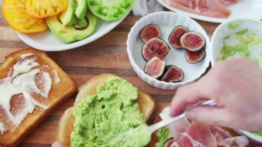 Bir kadın tost üzerine püre avokado yayılır ve hatıra domates dilimleri, prosciutto ve taze dereotu ekler 