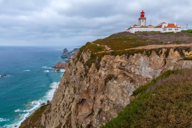 Deniz fenerinin manzaralı manzarası ve bulutlu bir gökyüzünün altındaki engin, turkuaz okyanusa bakan engebeli bir uçuruma tünemiş bir bina yığını.