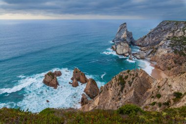 Praia da Ursa 'nın dramatik kıyı manzarası engebeli uçurumlar, turkuaz sular ve kasvetli bir gökyüzü altında dalgaları çarpıştırıyor.
