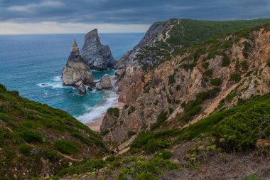 Praia da Ursa 'nın dramatik kıyı manzarası engebeli uçurumlar, turkuaz sular ve kasvetli bir gökyüzü altında dalgaları çarpıştırıyor.