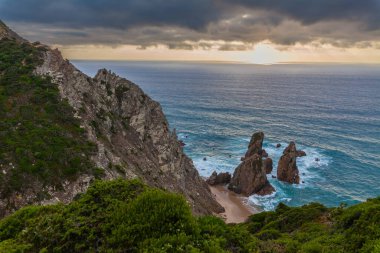 Praia da Ursa 'nın nefes kesici kıyı manzarası dramatik uçurumlar, turkuaz sular ve altın bir günbatımı gökyüzü altında deniz yığınları
