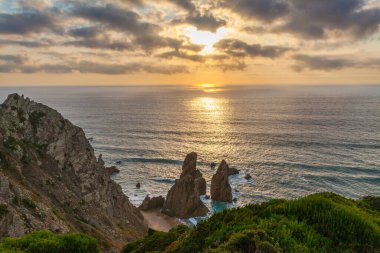 Praia da Ursa 'nın engebeli bir sahil şeridinde çarpıcı bir günbatımı dramatik deniz yığınları, sakin okyanus sularını yansıtan altın ışık, ve kayalıklardaki yemyeşil yeşillik