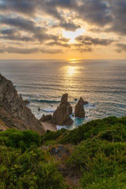 Praia da Ursa 'nın engebeli bir sahil şeridinde çarpıcı bir günbatımı dramatik deniz yığınları, sakin okyanus sularını yansıtan altın ışık, ve kayalıklardaki yemyeşil yeşillik