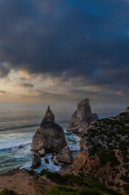 Alacakaranlıkta Praia da Ursa 'da dramatik bir kıyı sahnesi. Yükselen deniz yığınları, çarpan dalgalar ve yumuşak altın ışıkla parlayan kasvetli bir gökyüzü.