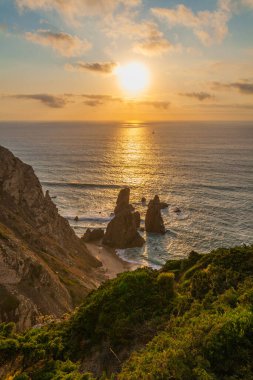Praia da Ursa üzerinde nefes kesici bir gün batımı engebeli bir kıyı şeridi okyanusa yansıyan altın ışıklarıyla dramatik deniz yığınları ve kayalıklarda yemyeşil bir yer.