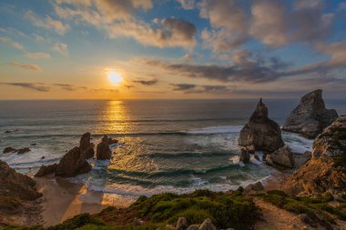 Praia da Ursa üzerinde nefes kesici bir gün batımı engebeli bir kıyı şeridi okyanusa yansıyan altın ışıklarıyla dramatik deniz yığınları ve kayalıklarda yemyeşil bir yer.