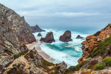Engebeli uçurumlar, tenha Praia da Ursa sahillerine çarpan turkuaz dalgalar ve yumuşak bir gökyüzünün altında dramatik deniz yığınları ile çarpıcı bir sahnedir.