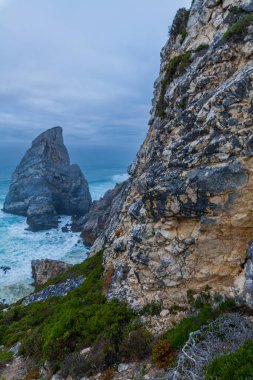 Praia da Ursa sahilindeki çalkantılı okyanus dalgalarından görkemli bir deniz yığını yükseliyor, yemyeşil ve kasvetli bir gökyüzünün altındaki engebeli kıyı şeridi ile çevrili.