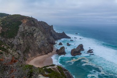 Praia da Ursa sahilindeki çalkantılı okyanus dalgalarından görkemli bir deniz yığını yükseliyor, yemyeşil ve kasvetli bir gökyüzünün altındaki engebeli kıyı şeridi ile çevrili.