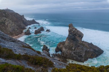Praia da Ursa sahilindeki çalkantılı okyanus dalgalarından görkemli bir deniz yığını yükseliyor, yemyeşil ve kasvetli bir gökyüzünün altındaki engebeli kıyı şeridi ile çevrili.