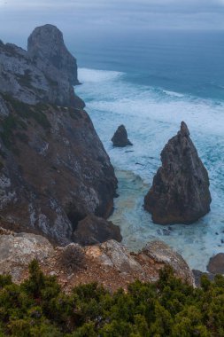 Praia da Ursa sahilindeki çalkantılı okyanus dalgalarından görkemli bir deniz yığını yükseliyor, yemyeşil ve kasvetli bir gökyüzünün altındaki engebeli kıyı şeridi ile çevrili.