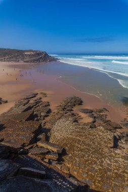 Turkuaz dalgaların kayalık bir sahile çarptığı çarpıcı bir sahil sahnesi, Apple sahilinde canlı mavi bir gökyüzünün altında kumlu kumsalla buluşması.