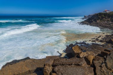 Turkuaz dalgaların kayalık bir sahile çarptığı, Praia das Mas 'ta canlı bir mavi gökyüzünün altında kumlu plajla karşılaşan çarpıcı bir sahile.