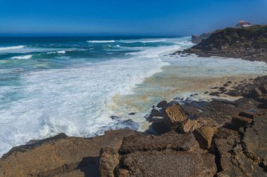 Turkuaz dalgaların kayalık bir sahile çarptığı, Praia das Mas 'ta canlı bir mavi gökyüzünün altında kumlu plajla karşılaşan çarpıcı bir sahile.