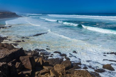 Turkuaz dalgaların kayalık bir sahile çarptığı çarpıcı bir sahil sahnesi, Apple sahilinde canlı mavi bir gökyüzünün altında kumlu kumsalla buluşması.
