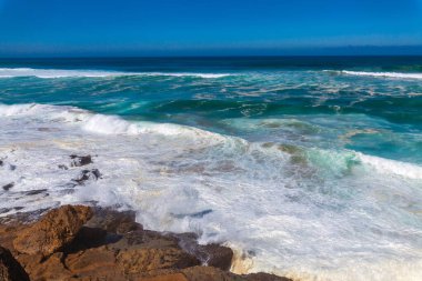 Güçlü dalgalar, dalgalı mavi gökyüzünün altında engebeli kıyı kayalarına çarpar. Okyanusa bakan uzak bir uçurum binası vardır.